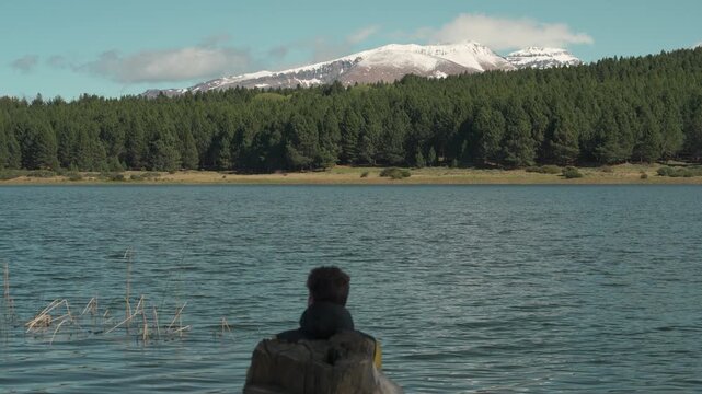 Man in a yellow jacket sitting on a bench overlooking the calm waters of Laguna La Zeta, with snow-capped mountain peaks and evergreen forests in the background at Esquel, Patagonia, Argentina.