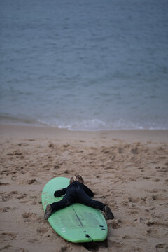 Little blonde girl in a black wetsuit lying on her stomach on a green surfboard on a sandy beach,