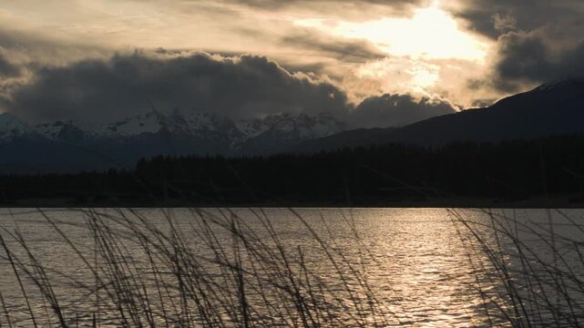 Golden hour light illuminating clouds over the snow-capped mountain range and silhouette of the pine forest at Laguna La Zeta, Esquel, Chubut, Argentina.