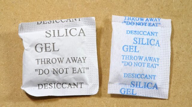 Two white silica gel desiccant packets with printed throw away and do not eat warnings on tan background, close-up video about moisture absorption, packaging safety and accidental ingestion prevention