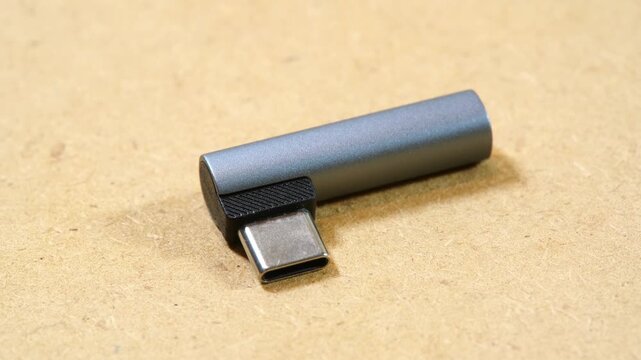 Small gray right-angle USB-C DAC audio adapter with exposed male connector on beige background, pocket digital-to-analog converter dongle for smartphones and devices without a headphone jack