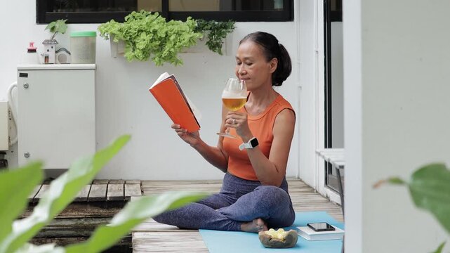 An elegant senior woman relaxing on her balcony, sitting on a yoga mat, reading an interesting book while holding a glass of craft beer in her peaceful backyard.