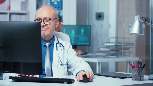 Elderly doctor works at a computer inside a glass walled private clinic, showing hospital professionals and daily medical office workflow.
