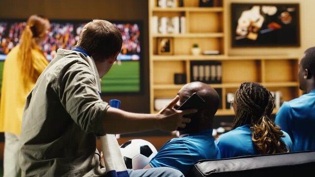 Man at home on couch holding mobile phone while watching match on television, sharing bad news with friends in apartment after seeing negative online match results during tense live game action.