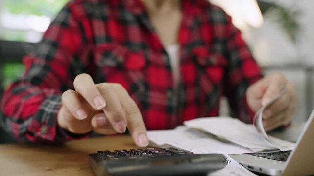 Closeup to hand of female accountant sat at desk with laptop computer using calculator to calculate company bills including wages utilities and taxes in office. Businesswoman working in her workplace.