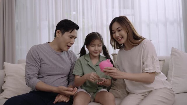 Happy Asian family saving money concept. Smiling father and mother with child girl sitting saving money into a pink piggy bank together for their daughter's future on sofa in the living room at home. 