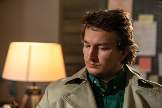 Young adult man in a trench coat sitting beside a warm lamp looking down in a moody noir detective atmosphere.