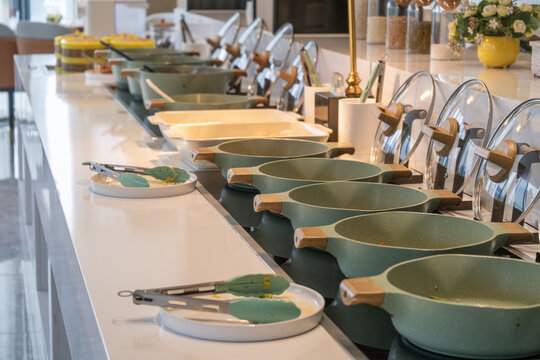 A contemporary hotel breakfast buffet station featuring a row of food pots sitting on an integrated induction cooktop to keep warm, along with serving tongs, offering a clean and basic presentation