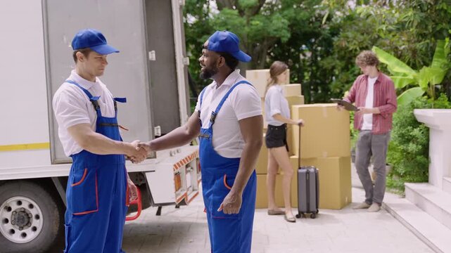 Happy professional movers in uniform standing talking and shaking hands after finishing unloading from a truck while a young couple of homeowners are checking and signing for delivery at a new home. 
