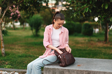 Fototapeta premium Woman sits on a park bench with a backpack, wearing a striped shirt and jeans, enjoying a quiet outdoor moment in a green park setting.