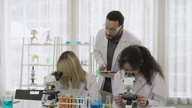 Two female students and a male science teacher wearing lab coats are using microscopes for learning and conducting chemistry experiments and liquid testing in a science class laboratory at school.