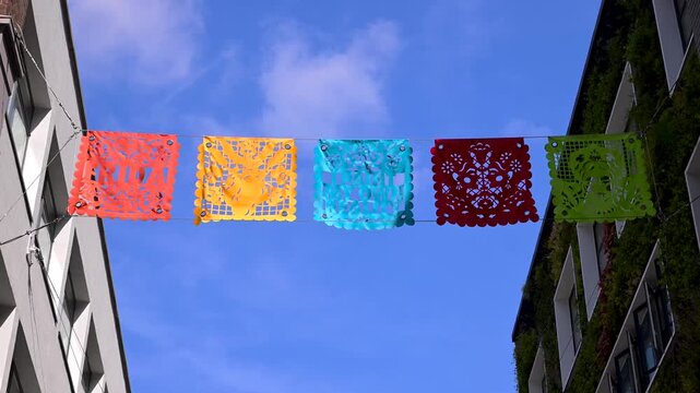 Bright papel picado flags in red, yellow, blue, teal and green hang between urban buildings against a blue sky.