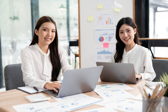 Two Confident Asian Businesswomen Smiling at Camera While Working on Laptops