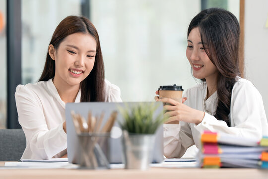 Two Happy Asian Businesswomen Having Coffee and Working on Laptop Together