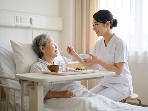 病床の高齢女性に優しく声をかけながらご飯を食べさせる看護師の女性