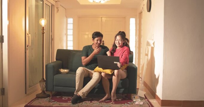 Asian woman, asian man lover have good news, video call, happy asia couple with laptop celebrating on sofa, hug and excited in living room. Happy asian couple checking online for honeymoon travel. 