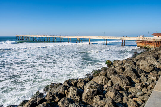 wide-angle view of churning white ocean surf crashing against a rocky shoreline next to a long public pier under a clear blue sky