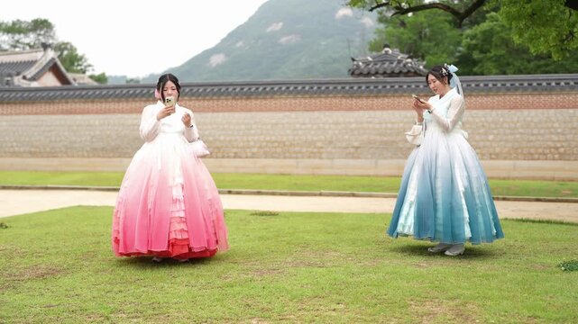 Two women in colorful hanboks dance gracefully on green grass. They move with elegance, hands clasped near their chests. Traditional Korean architecture frames the scene behind them
