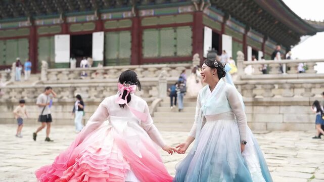 Two women in flowing hanboks dance joyfully. Their smiles reflect pure delight and connection. They hold hands, moving gracefully together. Behind them, ancient temple architecture stands grand