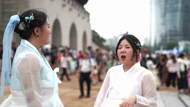 Two women in traditional Korean attire converse outdoors. One wears light blue, the other pink and white. They stand near an arched stone structure. Background shows city buildings and pedestrians
