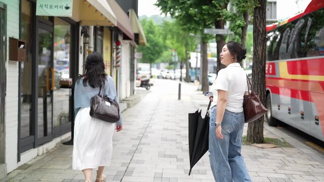 Two women stroll down a tree-lined sidewalk. One carries an umbrella, the other walks bare-handed. They pause near a bus, chatting casually. Street shops line the pavement behind them