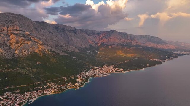 Brela Croatia shows a coastal area with buildings, green land, and mountains. The sea meets the shore. Clouds are present in the sky. Time of day is not specified.