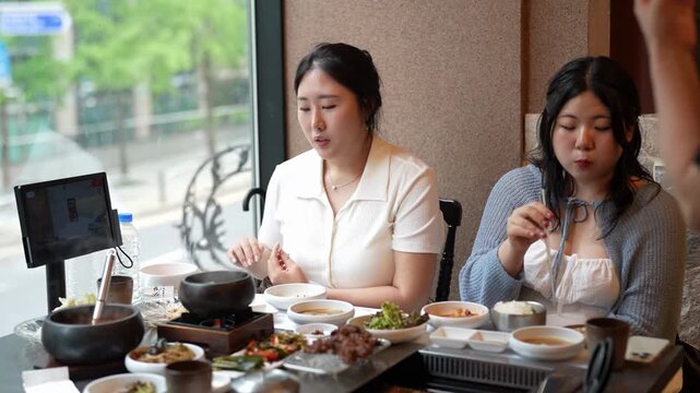 Two women enjoy a shared Korean meal together. They gesture, speak, and eat with relaxed, happy expressions. Hot pot sits at the center of their table. The scene suggests casual