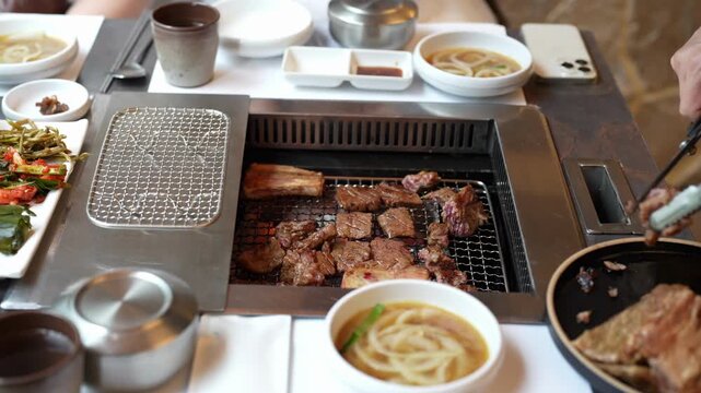 Person grills meat over built-in tabletop BBQ. Dipping noodles in warm broth adds flavor. Hands show care and focus during cooking. Great for restaurant or food culture scenes