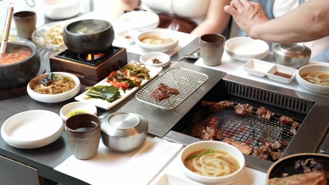People enjoy grilled meat with side dishes at a table. Hands tend to sizzling grill and steaming bowls. Warm lighting highlights communal dining joy. Perfect for restaurant, food, or culture content