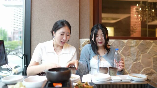 Two women smile at hot pot dinner. One holds a bottle, the other gestures with chopsticks. They sit beside a warm, glowing pot. Light streams through a window behind them. The scene feels cozy