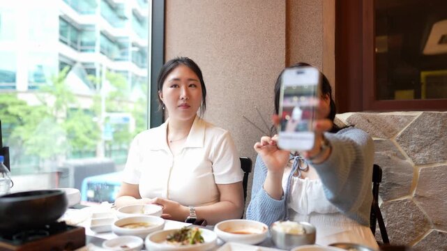Two women dine together at a cozy restaurant. One takes a photo, the other smiles and eats. They sit at a table filled with dishes and a pot. Warm lighting, stone walls. Perfect for social, lifestyle