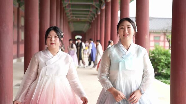 Two women in hanbok walk side by side. They smile gently, enjoying the moment. Traditional architecture frames their journey. Others in colorful attire stroll behind them