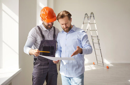 Young man standing with cheerful male foreman in safety helmet with blueprint in hands in empty new apartment discussing design project or repair renovation. Real estate and moving concept