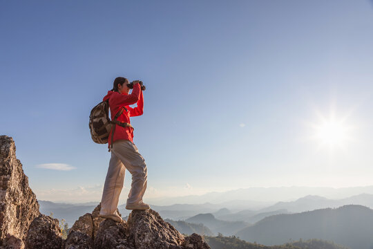 Asian girl hiker with backpack holding binoculars standing on top of the rock mountain sunset sky background
