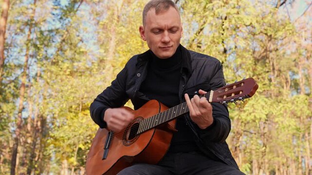 Portrait of smiling caucasian artist plays wooden acoustic guitar in autumnal park. Young songwriter plays string musical instrument hobby outside in nature fall time. Audio music healing in natural