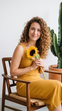 Smiling young woman with curly hair sitting on a wooden chair holding a bright yellow sunflower in a minimalist interior setting with a large cactus