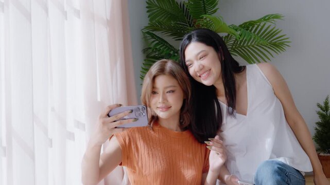 Two young Asian women smiling and happy while taking a selfie in their living room at home, video call, meeting conference, looking at the camera