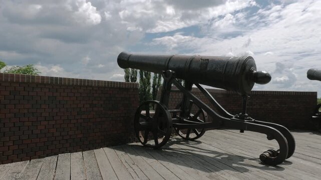Historic cast-iron cannon on a cast-iron carriage atop a wooden fortress platform in Chernihiv, Ukraine, with a brick wall in the foreground and a sunny, cloud-filled sky behind.