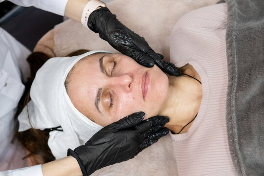 Facial massage. Cosmetician occupation. Woman professional beauty master in black gloves using product for treatment in spa salon.