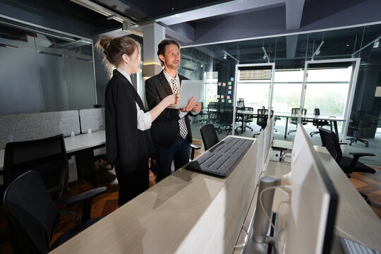 Young businesswoman with a bun holds a tablet while her dark-haired colleague looks toward the screen, the two standing among desks in a sunlit modern coworking office.