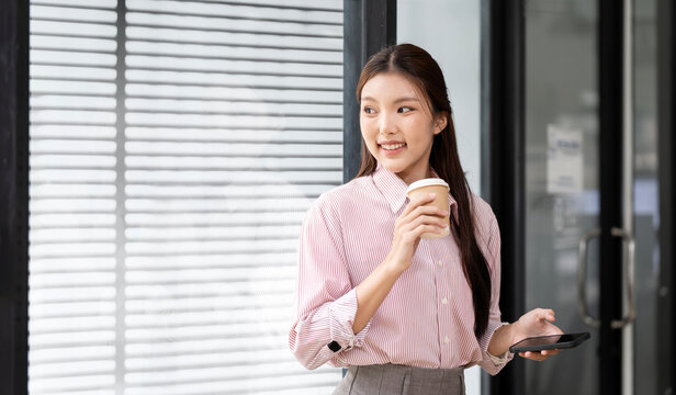 Happy Young Asian Businesswoman Holding Coffee Cup and Smartphone in Office