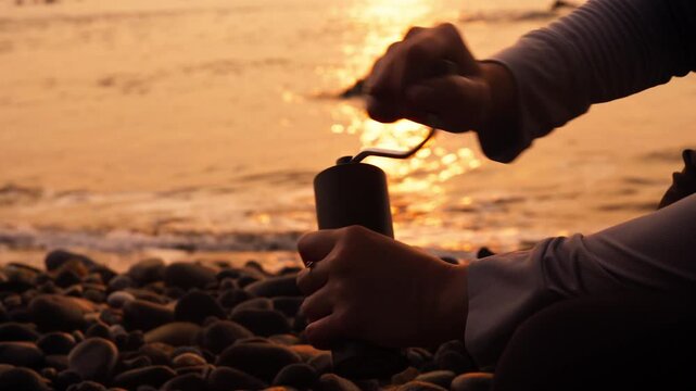 a person grind coffee bean using manual grinder in golden sun sets over pebbled shore