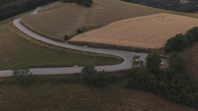 Drone view of a winding rural road curving through rolling farmland bordered by golden wheat fields and green vegetation.