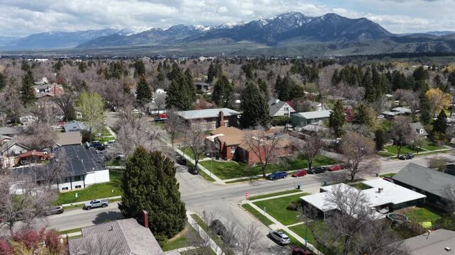 Snow-capped mountains rise above quiet Bozeman, Montana neighborhood, bare early spring trees surrounding brick houses plus parked cars lining shaded residential streets. Aerial drone wide shot
