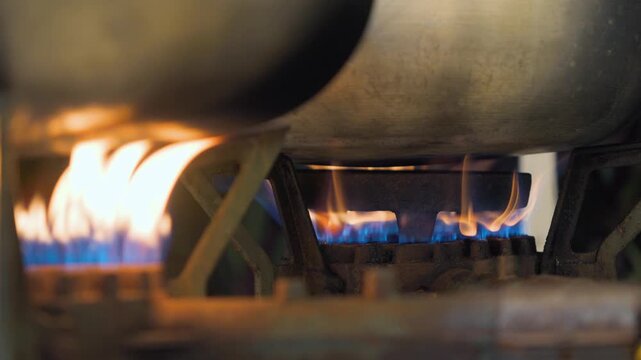 A close-up shot of industrial gas stove burners with visible blue flames heating large metal pots, demonstrating a professional kitchen cooking process