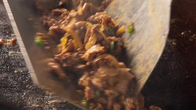 Sizzling meat sizzles in a flat top hot griddle with onions and green chilies. Spatula flips the mixture, releasing steam and aroma