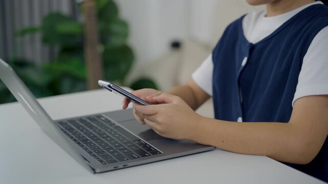 Child using smartphone while sitting at desk with laptop computer in modern home interior focusing on electronic device communication and technology concept