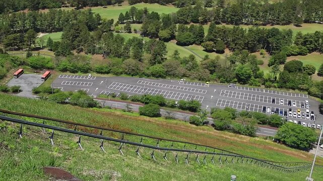 Panoramic horizontal footage from the funicular on Mount Omuro in Izu, Japan, overlooking lush green slopes, parking area, trees and distant sea horizon. Beautiful sightseeing and nature travel scene.