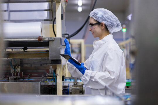 female worker inspecting manufacturing factory production line using digital tablet while wearing safety clean room uniform hairnet and blue protective glove showing attentive look