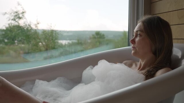 Lost in thought, a woman enjoys a delightful bubble bath within a modern bathroom, her gaze fixed on the expansive lake and green wilderness visible through the window on a peaceful day.